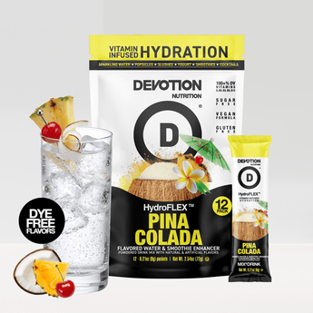 A Devotion Nutrition HydroFlex Pina Colada flavored powder bag stands next to a glass of iced water with pineapple cherry and lime garnish plus a single stick pack. The packaging is white black and yellow with fruit and coconut graphics.