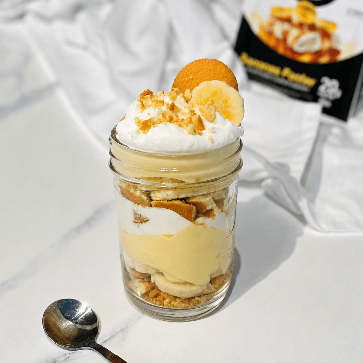 A layered dessert parfait in a clear glass mason jar, topped with whipped cream, banana slices, and a banana pie piece, set on a white marble-like surface with a spoon and a blurred product box in the background.