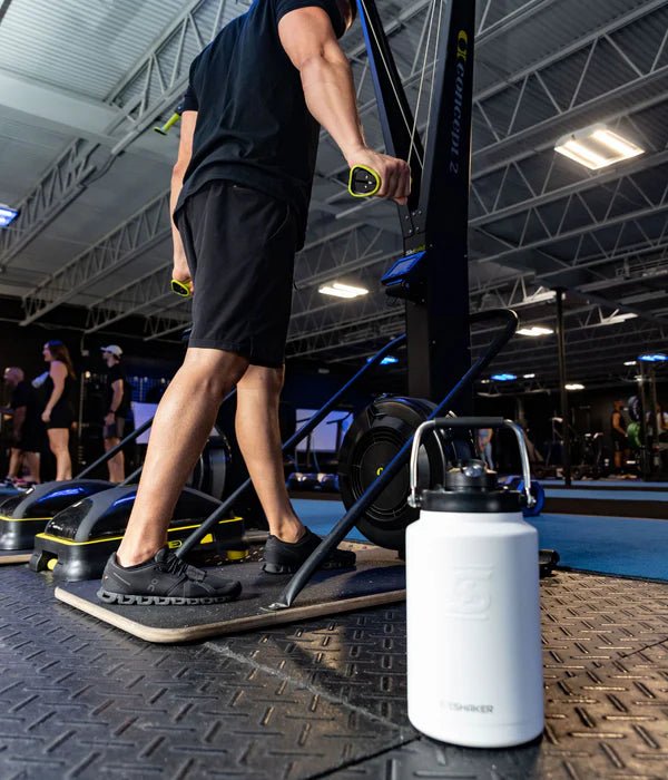 Person using exercise equipment in a gym with a white water bottle in the foreground.